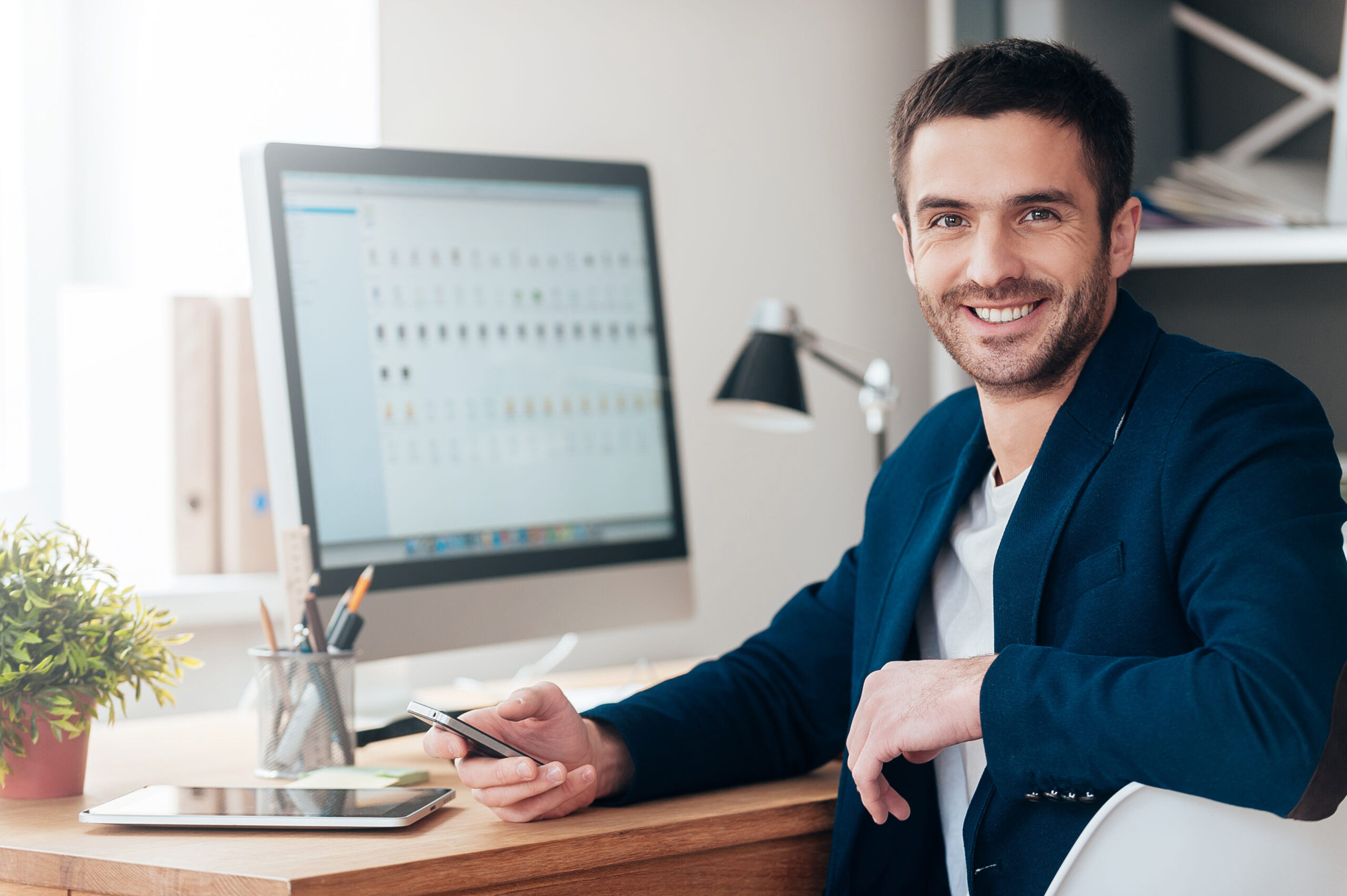 Confident and successful. Confident young man holding smart phone and smiling while sitting at his working place in office
